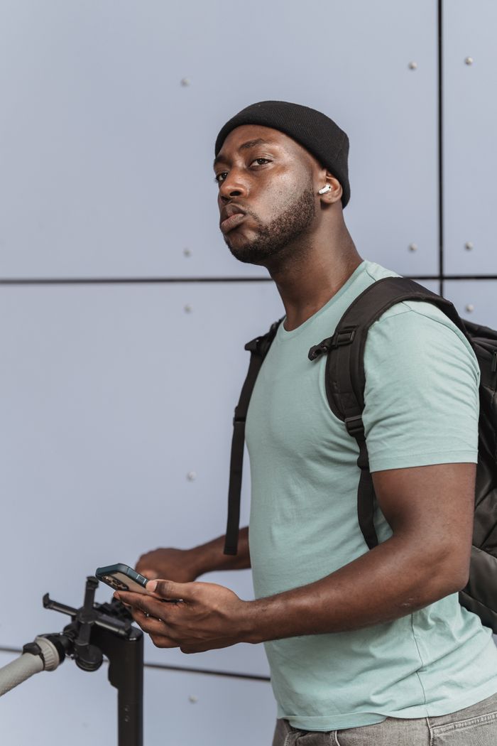 A man standing with an electric scooter holding a smartphone, wearing a backpack and beanie, looking thoughtful.